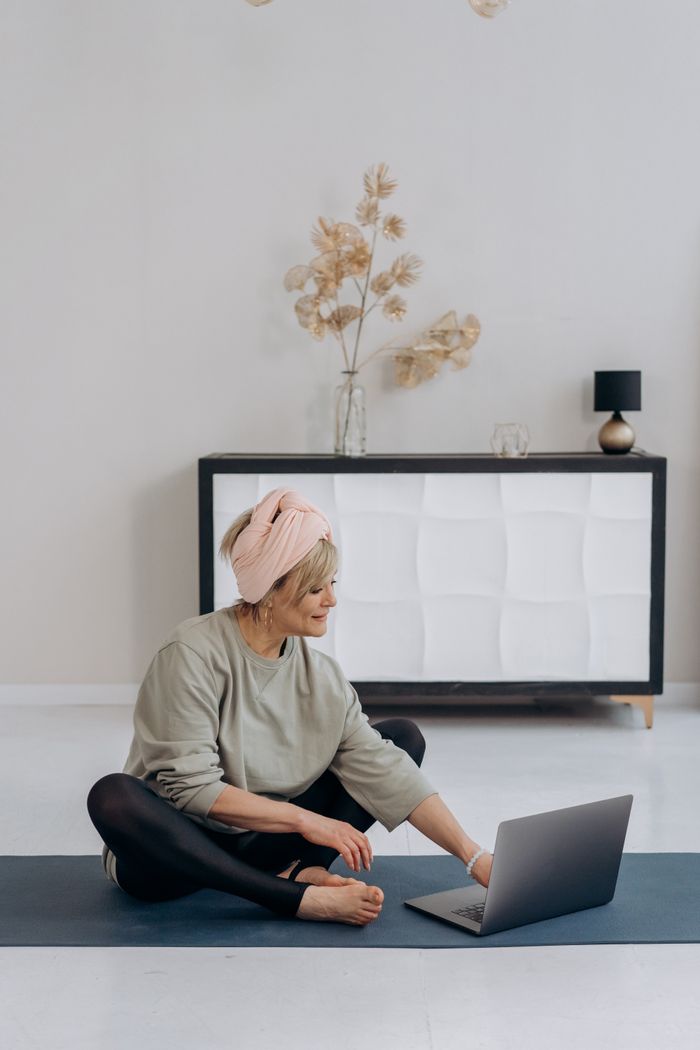 our-story Elderly woman sitting on a yoga mat using a laptop in a stylish living room.